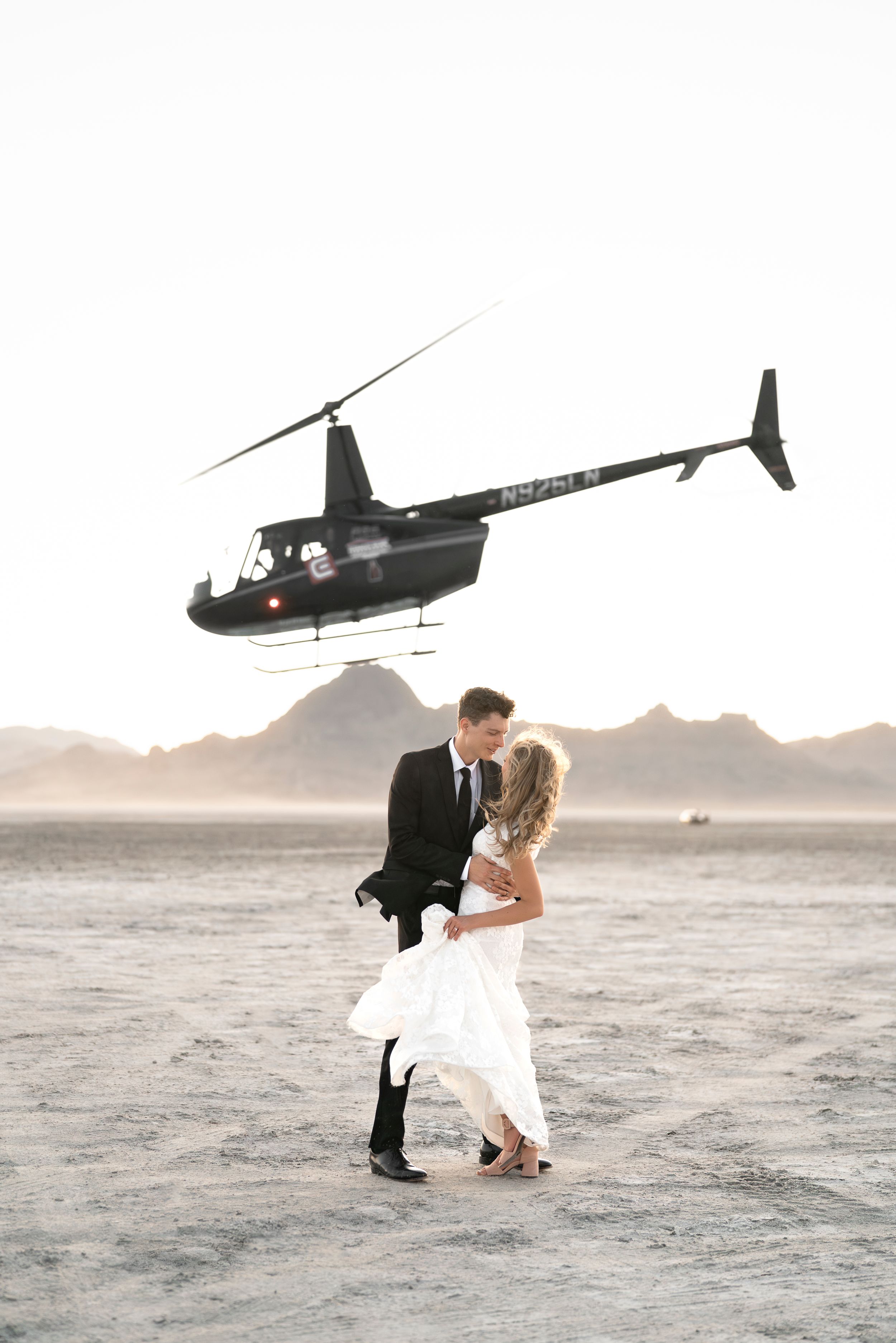 Wedding couple dancing beneath a hovering helicopter on a desert landscape with mountains in the distance.