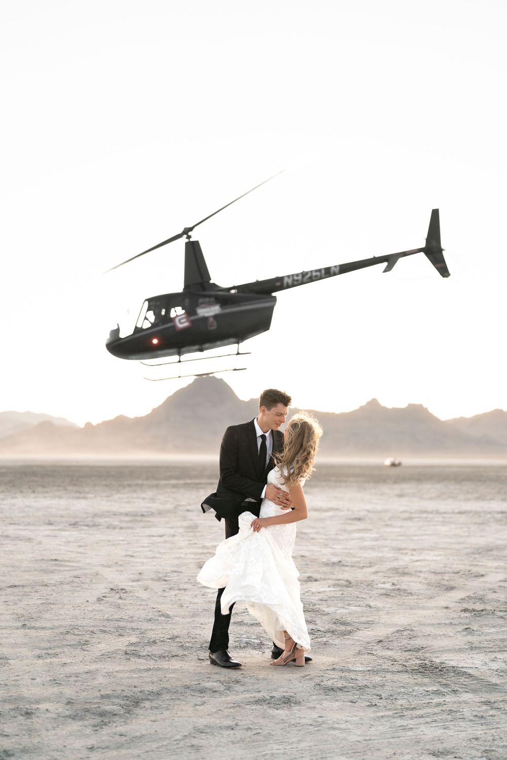 Wedding couple dancing beneath a hovering helicopter on a desert landscape with mountains in the distance.