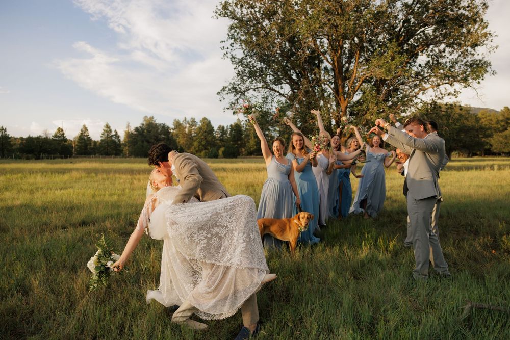 Wedding party celebrating in a sunlit green field with bridesmaids in blue dresses and groomsmen cheering.