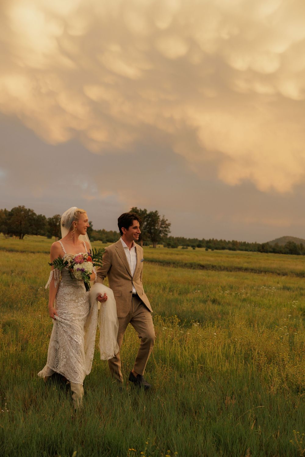 Bride and groom walking hand in hand through a lush green meadow under stormy golden clouds.