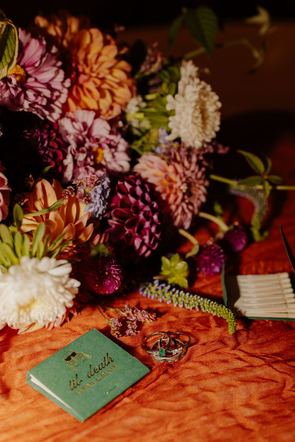 Colorful dahlia wedding bouquet with rings and a green matchbook on an orange textured surface.