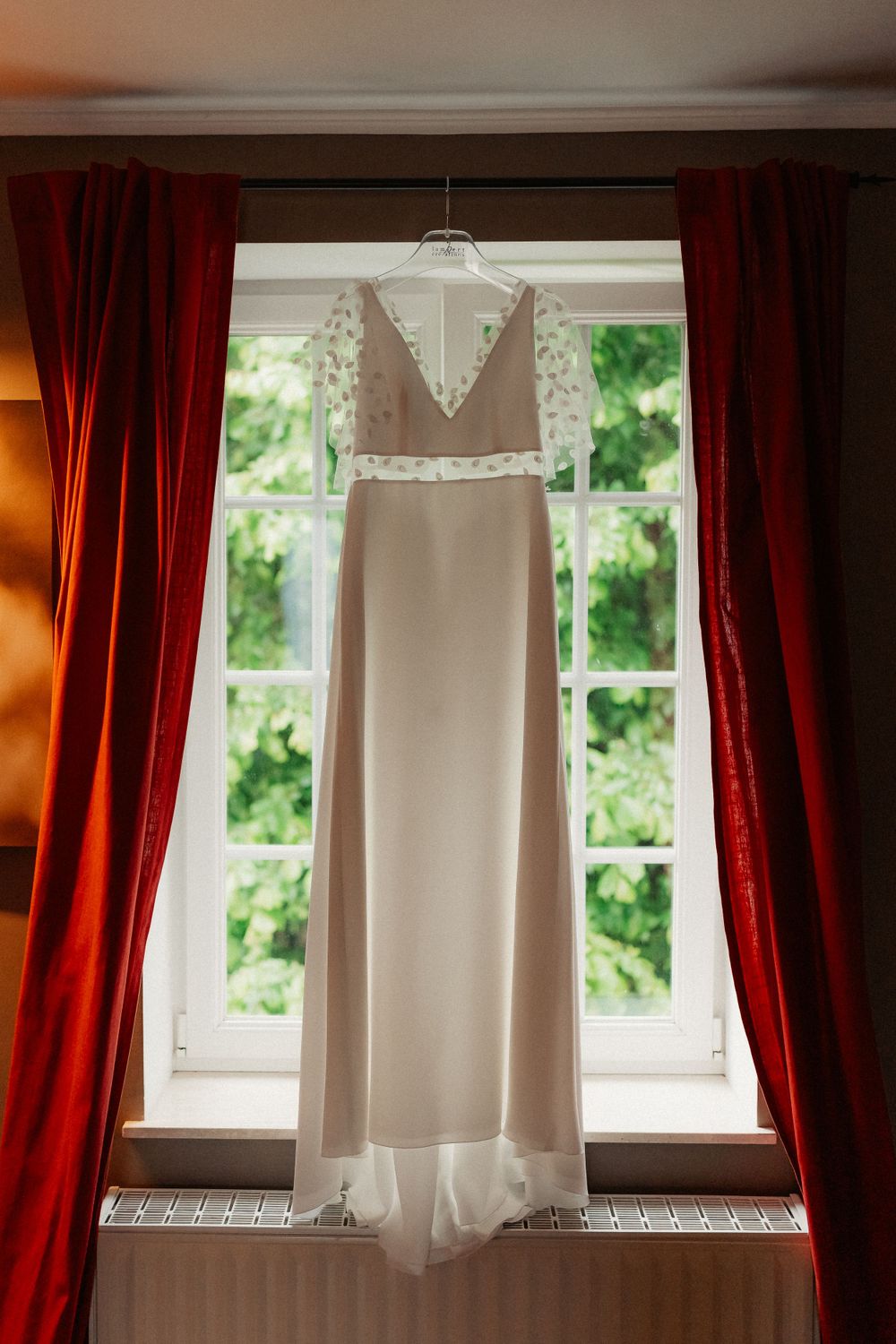 Elegant white wedding dress hanging in window frame with red velvet curtains.