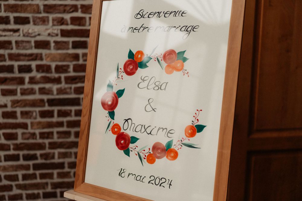 A wedding sign decorated with watercolor floral designs in orange and pink tones against a brick wall.