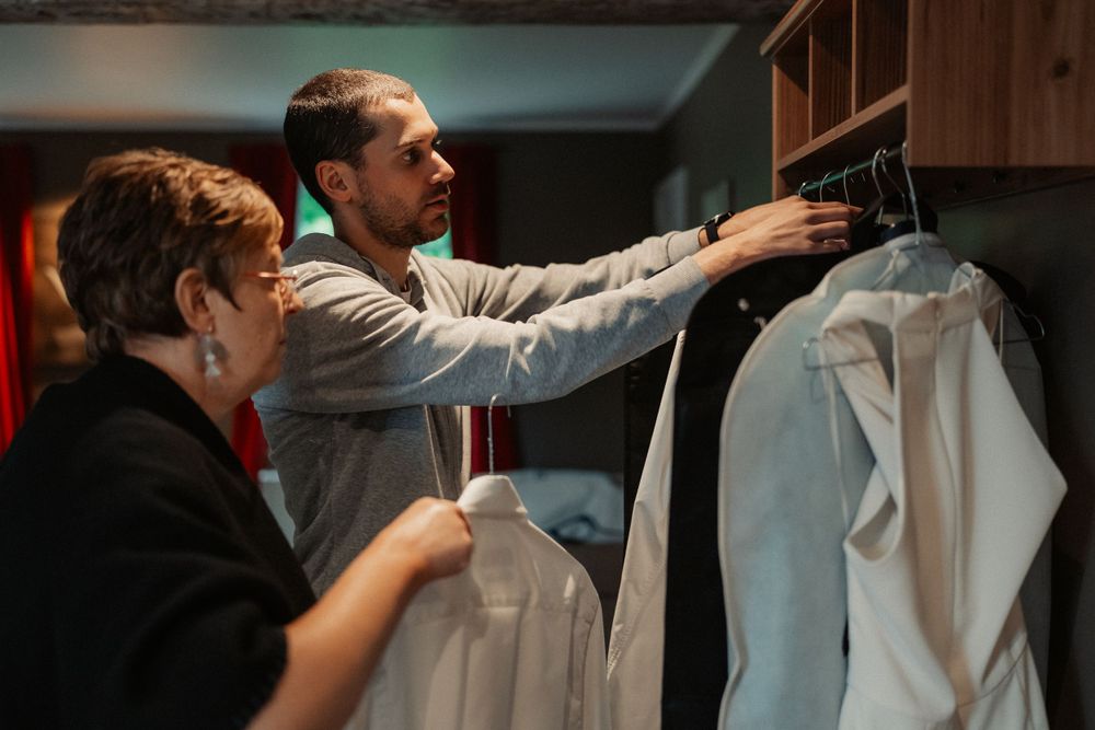 Two people selecting formal attire from a closet with wooden shelving.