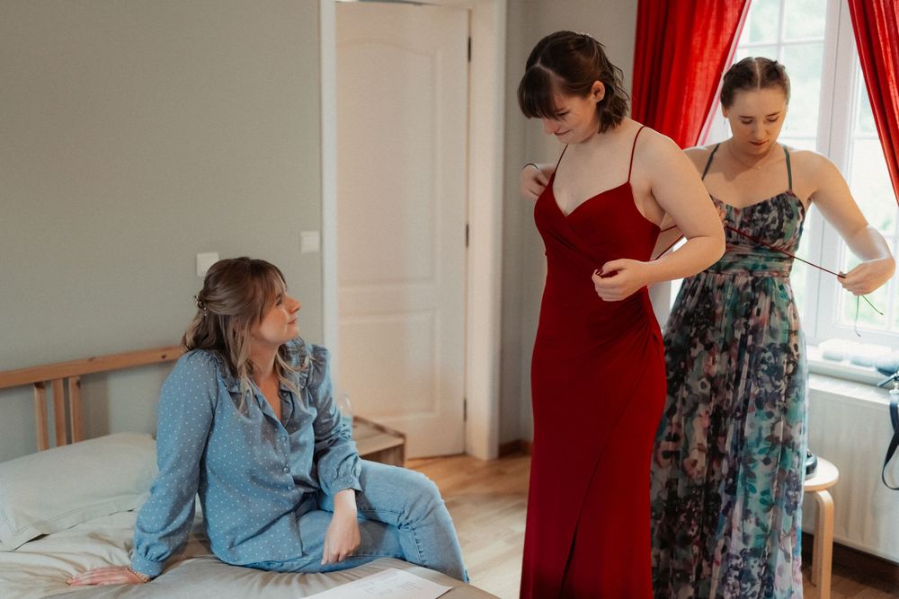 People in formal dresses gathered in a bedroom with red curtains.