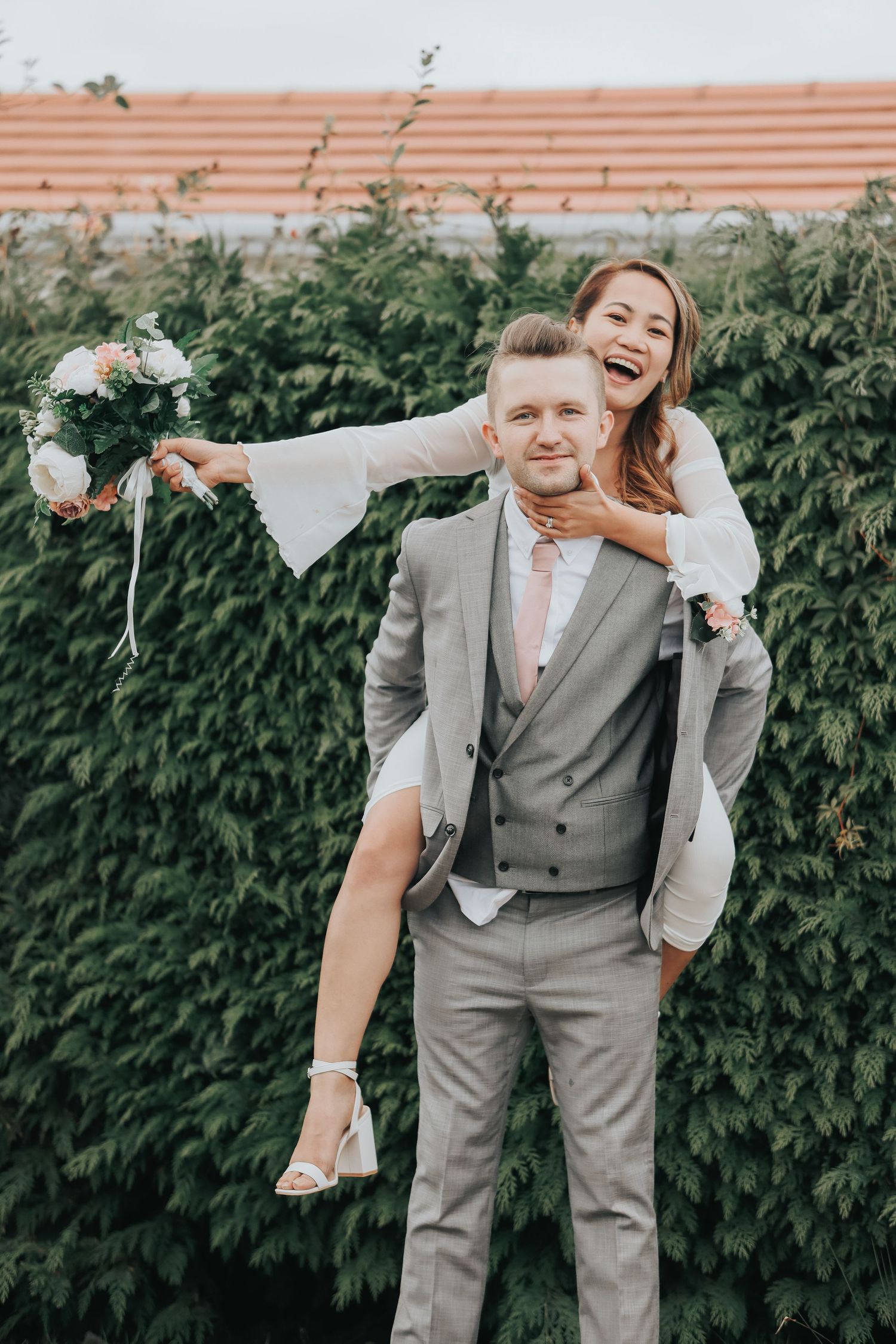 Playful couple poses against green hedge backdrop during outdoor wedding photoshoot.