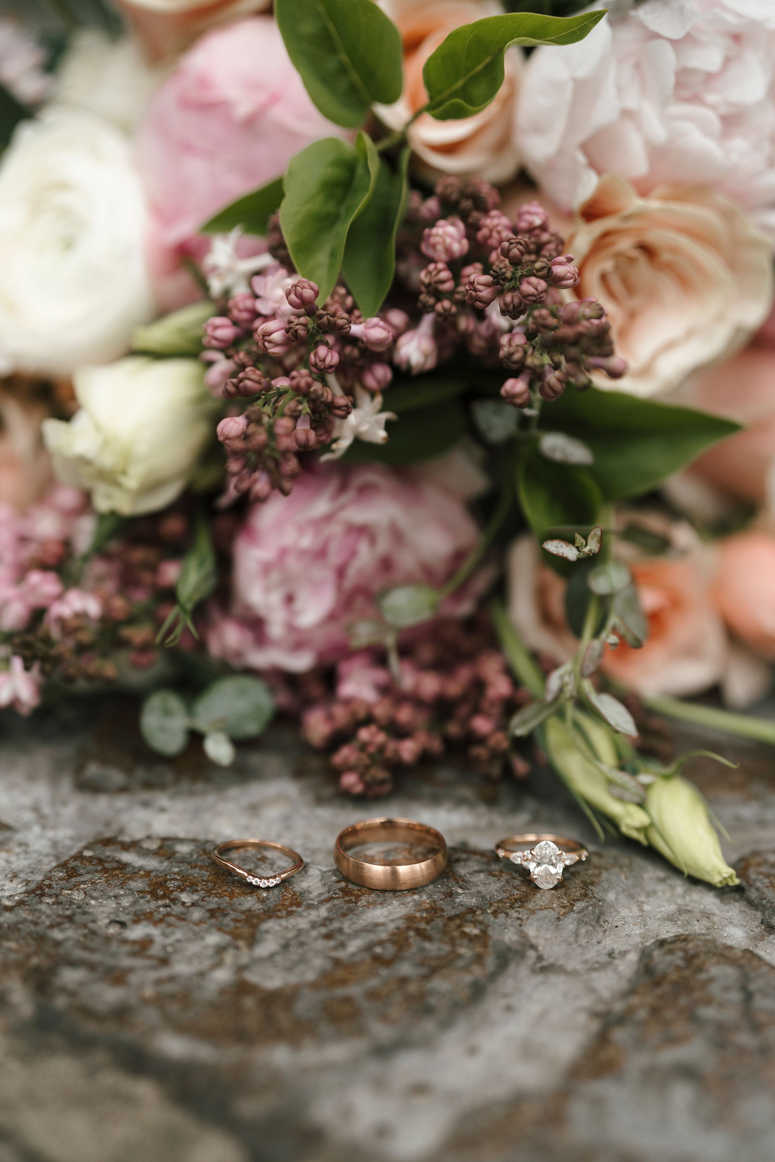 Wedding rings displayed on stone surface with soft pastel floral arrangement featuring roses and greenery in background.