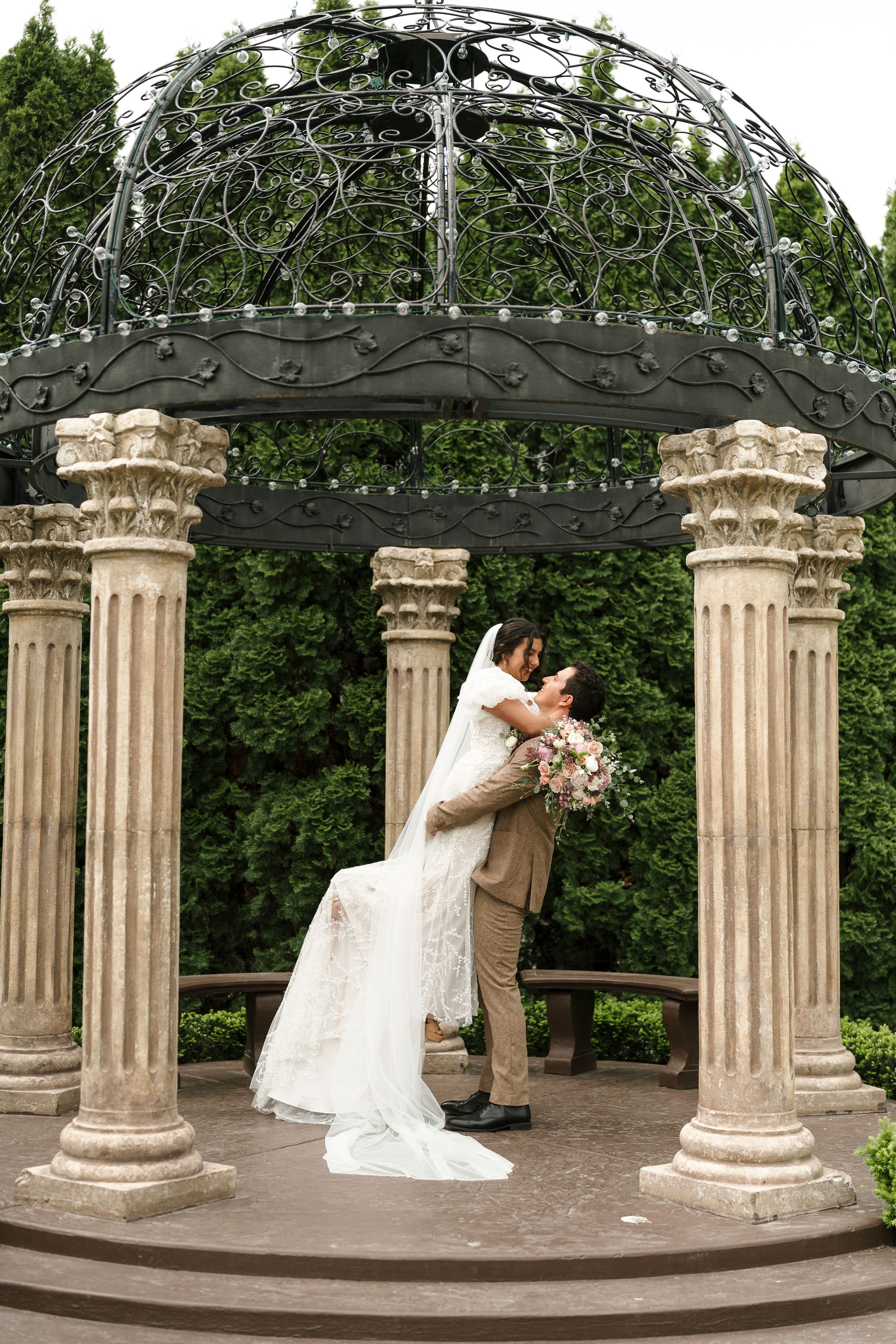A romantic moment captured under an ornate iron gazebo with classic Corinthian columns in a garden setting.