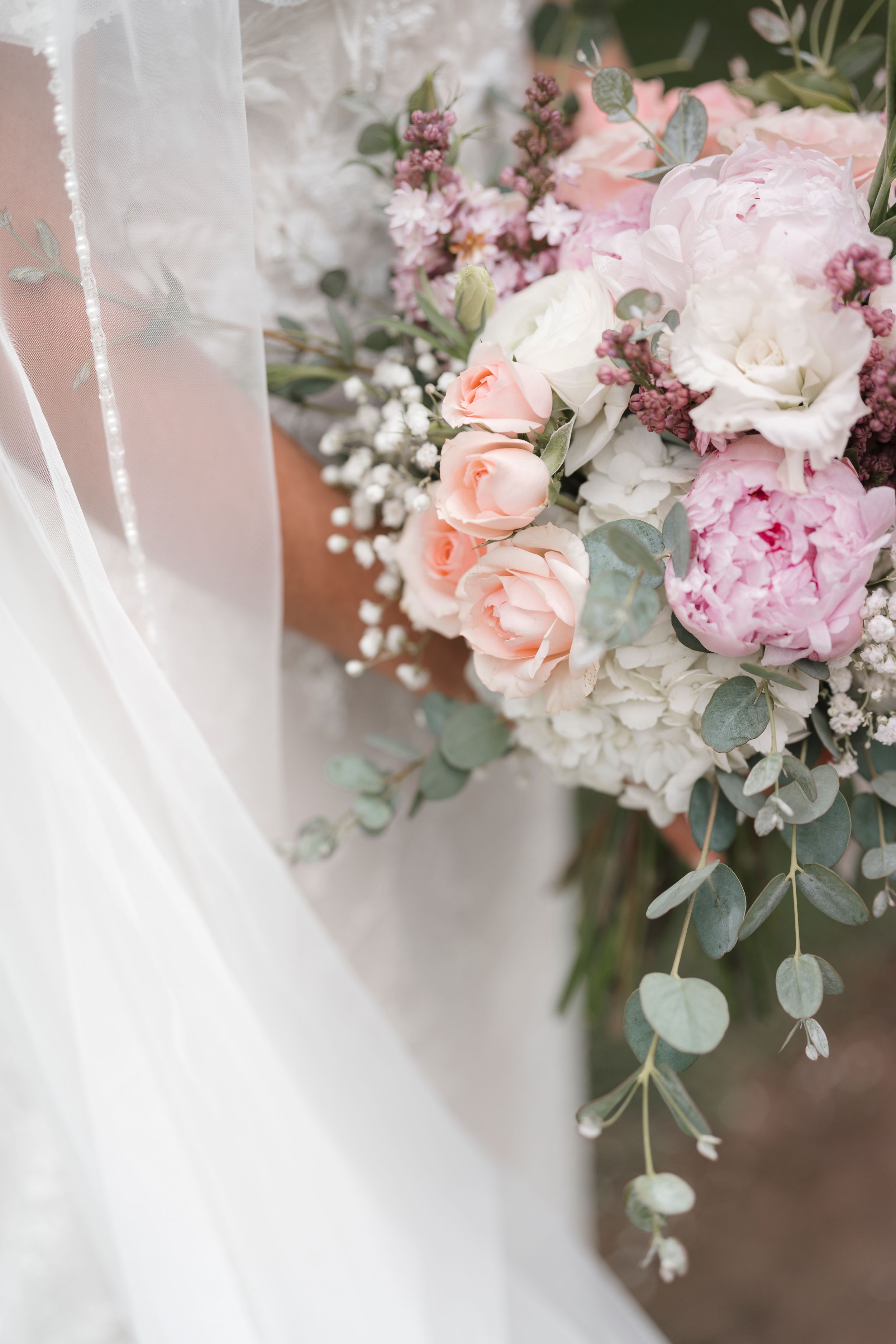 A delicate bridal bouquet featuring soft pink roses, peonies and eucalyptus against flowing white tulle.