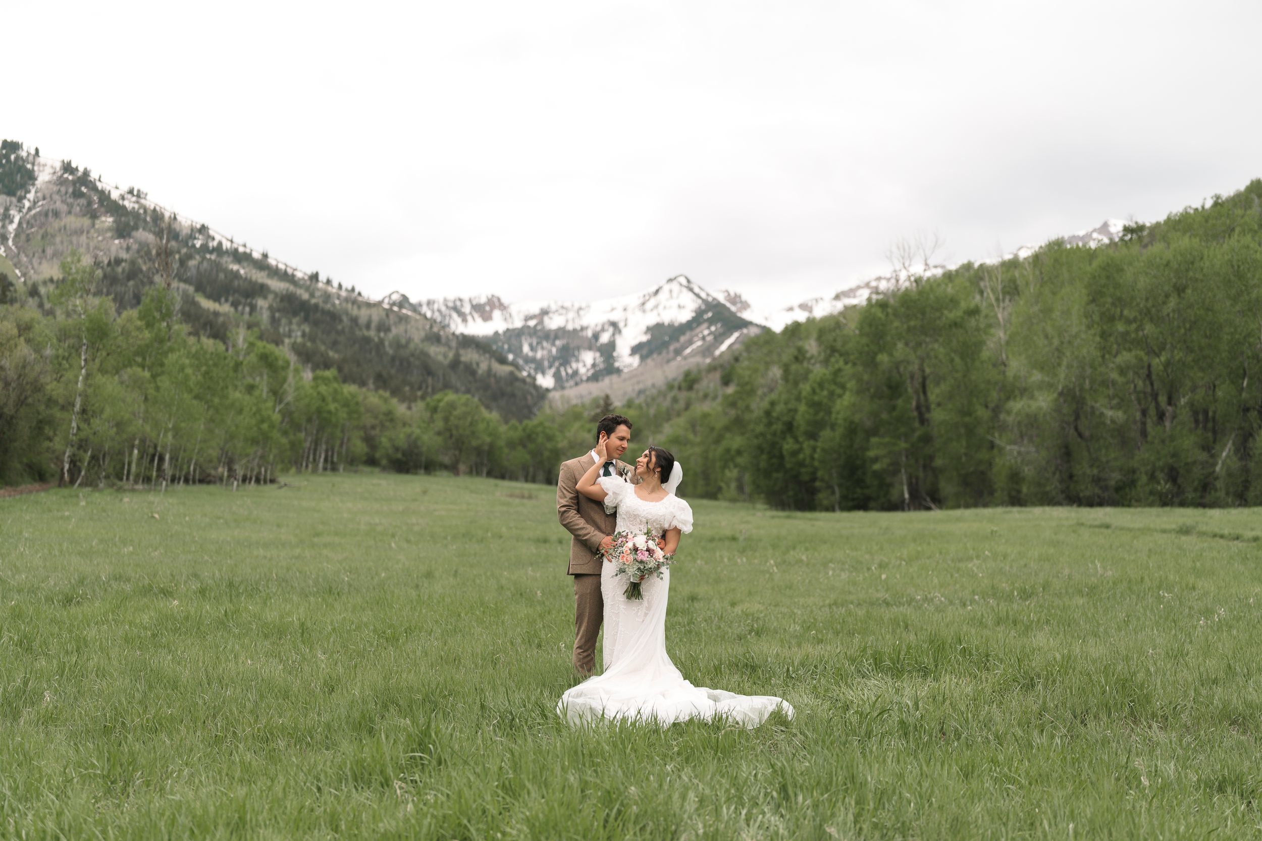 A scenic mountain wedding portrait in a green meadow with snow-capped peaks and aspen trees in the background.