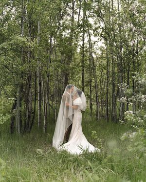 A romantic wedding portrait in a lush forest setting with tall trees and wildflowers surrounding the couple in soft light.