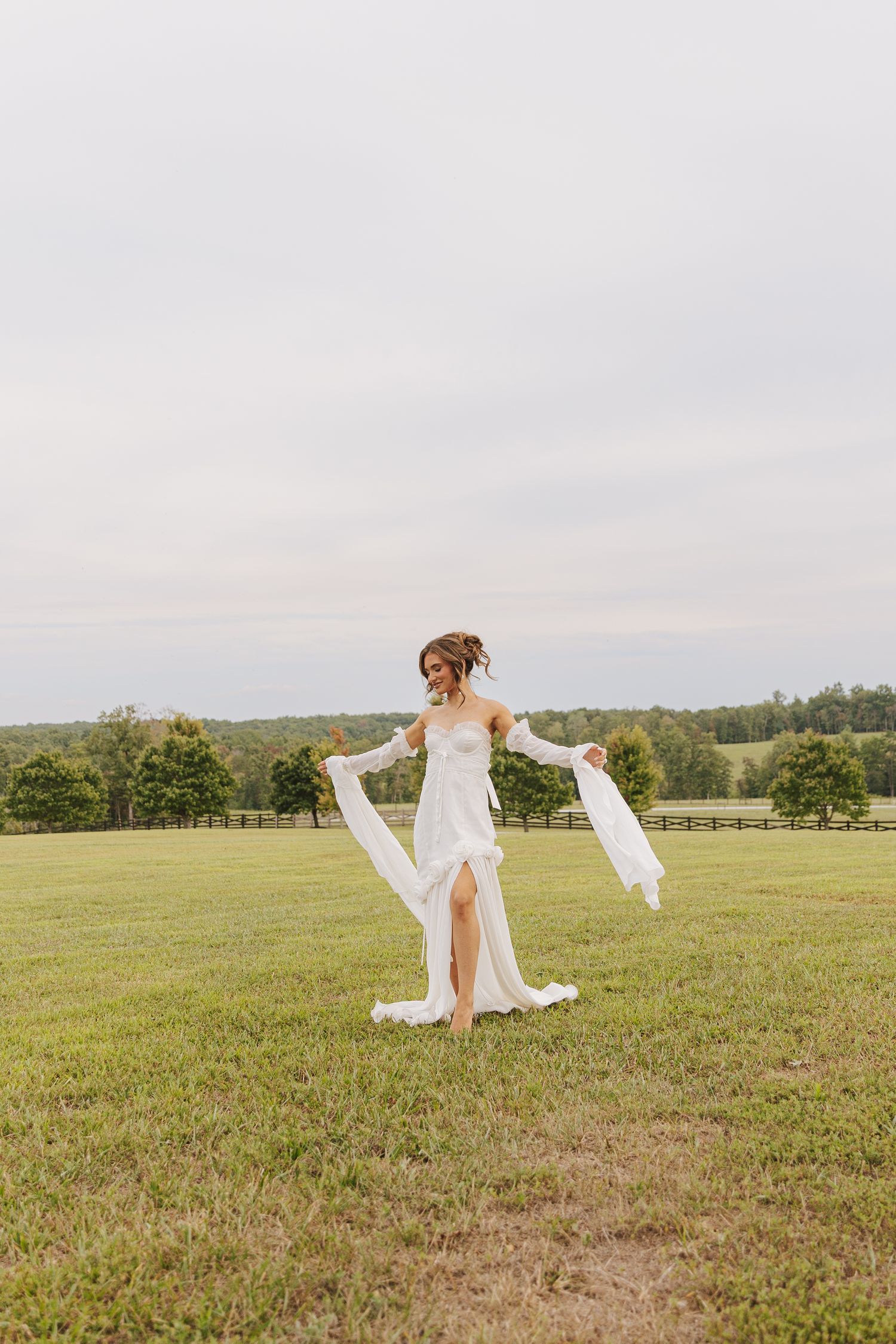 A person in a white flowing dress stands on a grassy hilltop with trees in the distance on an overcast day.