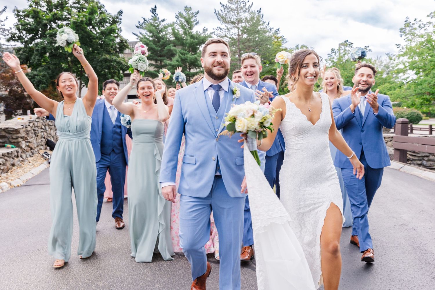 Celebratory wedding party walking outdoors in light blue and white formal attire.