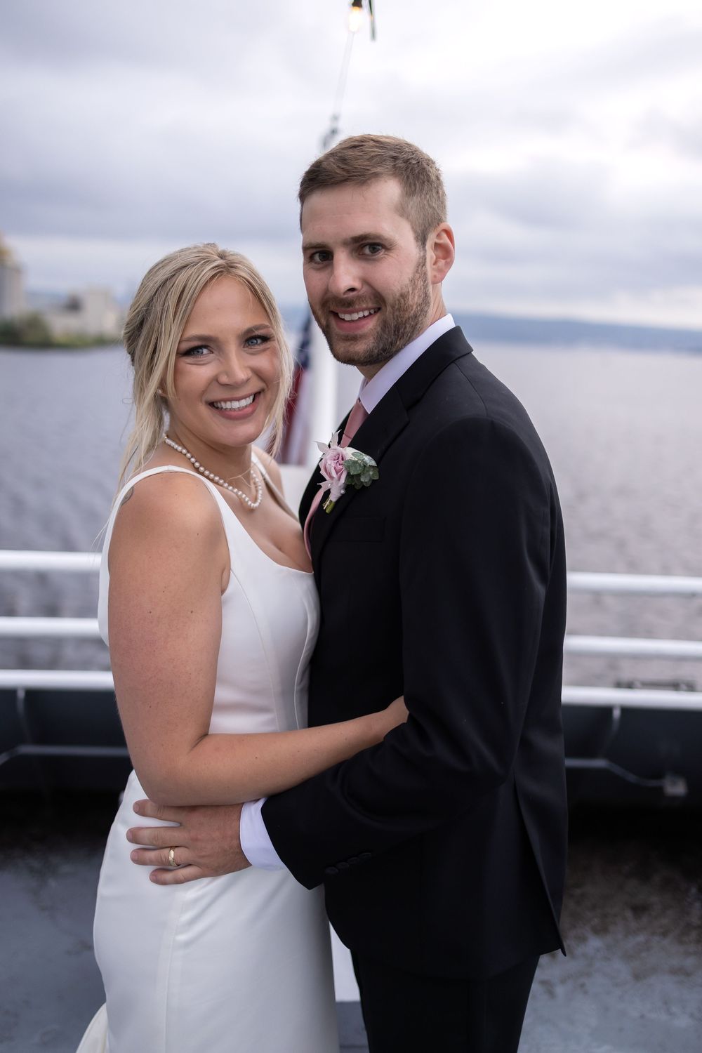 A newlywed couple embraces on a boat deck overlooking water with cloudy skies in the background.