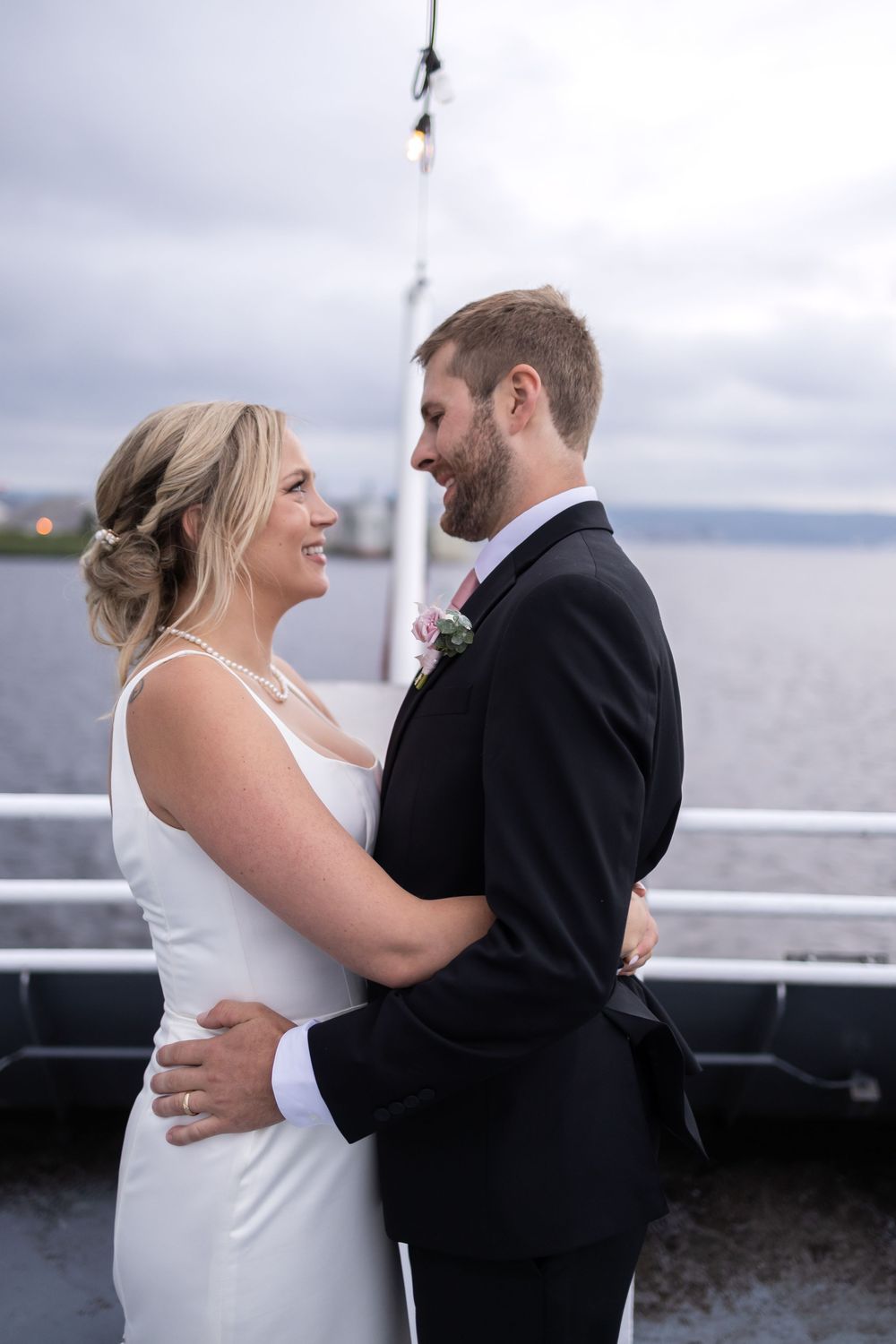Newlyweds share a joyful moment while embracing on a boat deck with water and cloudy skies in the background.