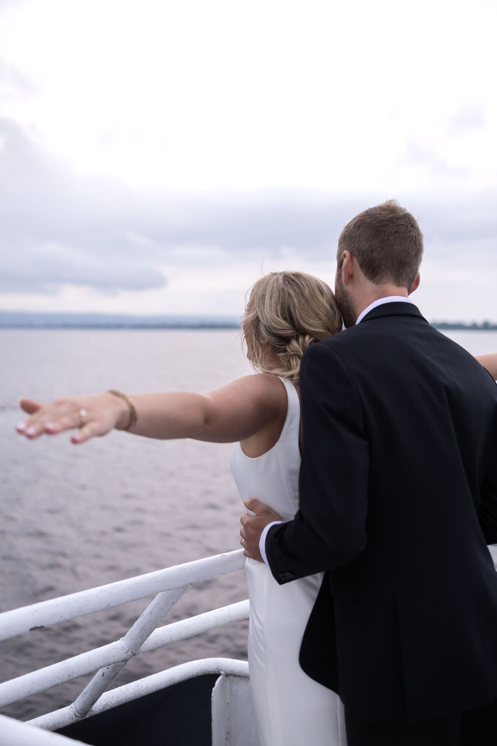 A couple in wedding attire stands together on a boat deck overlooking a misty waterfront view.