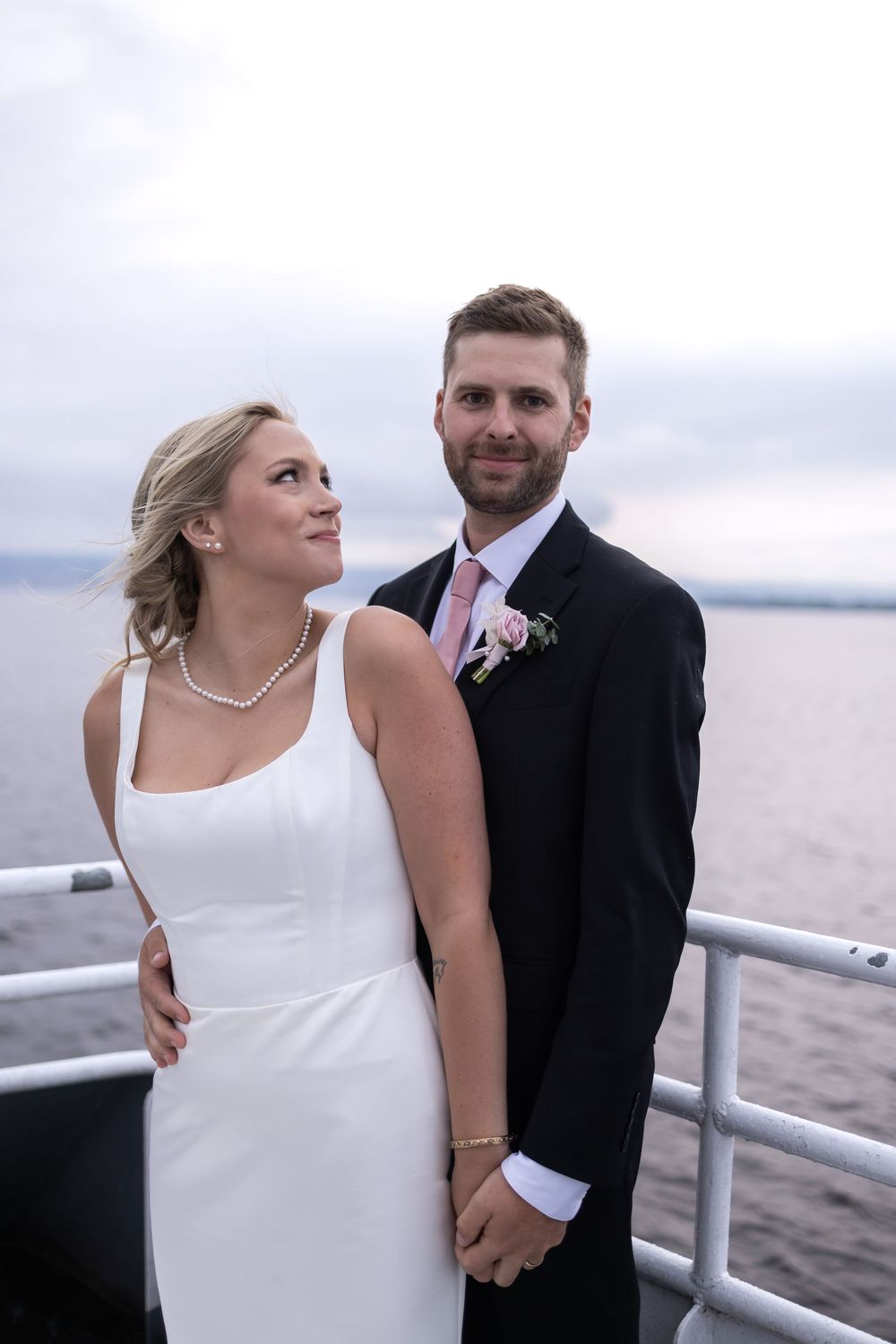 A couple in wedding attire share an intimate moment together on a boat deck overlooking calm waters on an overcast day.