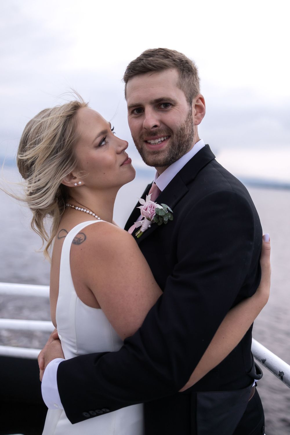 A romantic wedding couple embraces on a deck overlooking water, with the bride in white and groom in formal black attire.