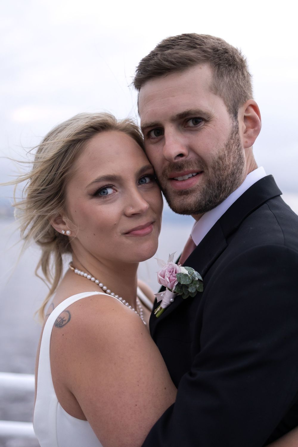 A bride in white dress with pearl necklace and groom in black suit embrace during outdoor wedding photo shoot.