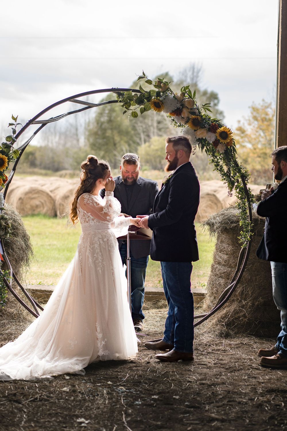 A rustic outdoor wedding ceremony under a circular floral arch with hay bales in the background on a cloudy day.