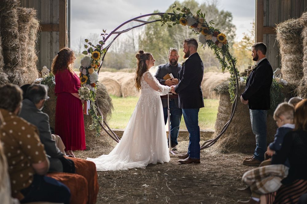 Couple stands beneath rustic floral arch during intimate barn wedding ceremony with guests seated on hay bales.