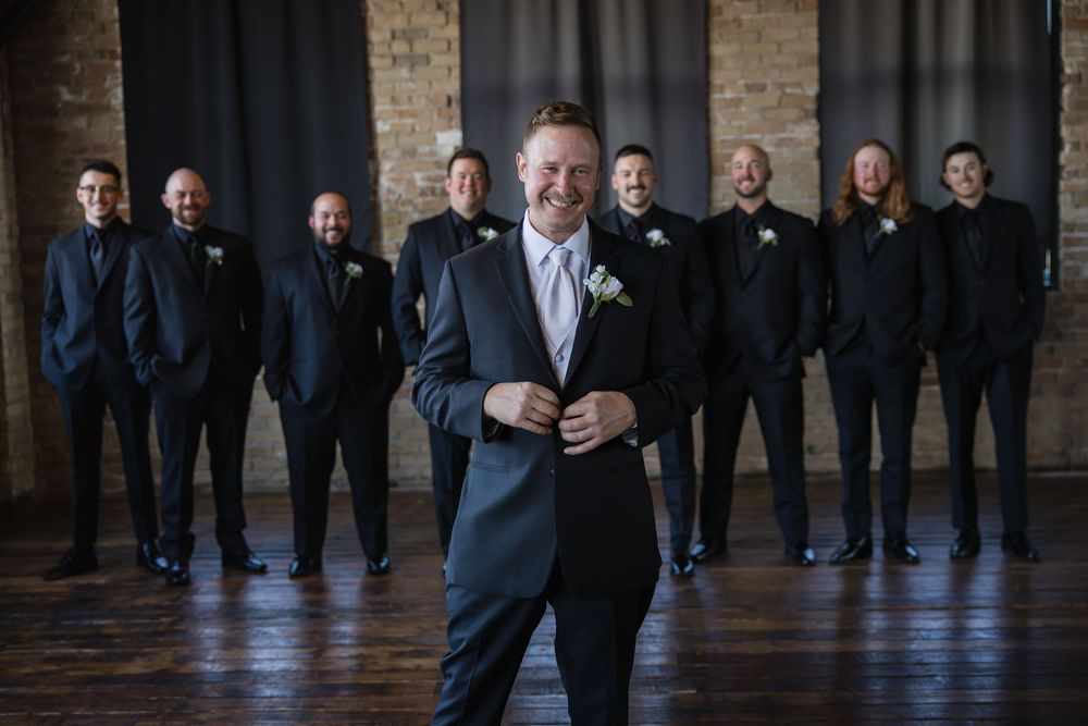 A groom in a navy suit stands confidently in front of his groomsmen wearing black suits in an industrial brick venue.