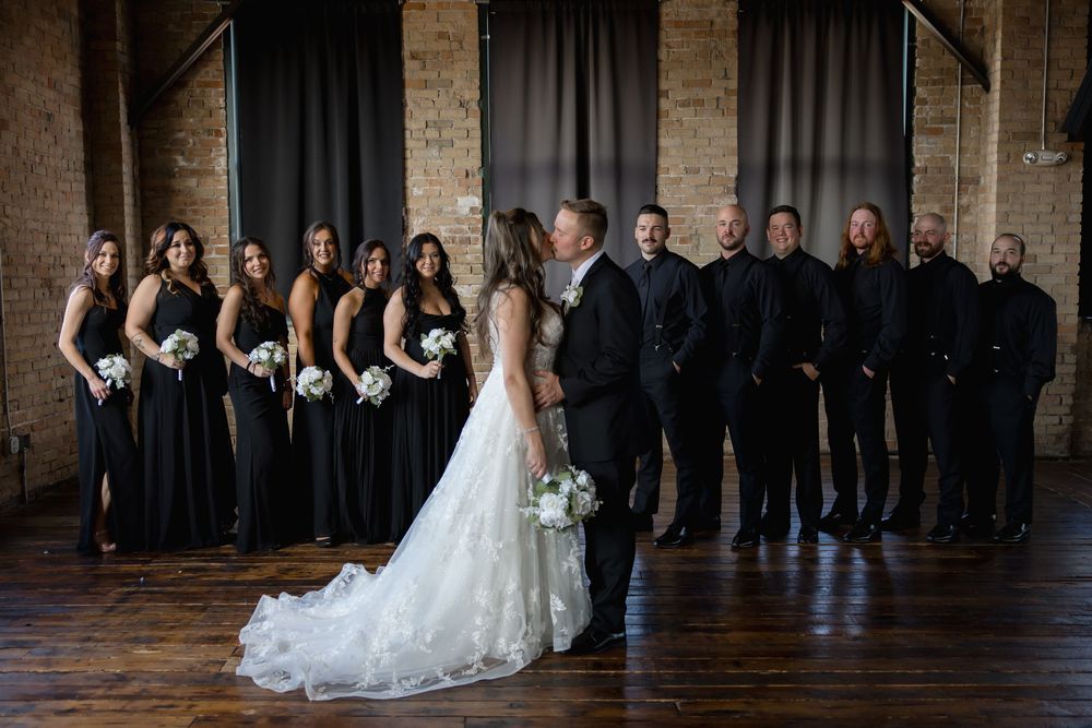 Wedding party lined up in an elegant brick venue with bridesmaids in black dresses and groomsmen in matching suits.