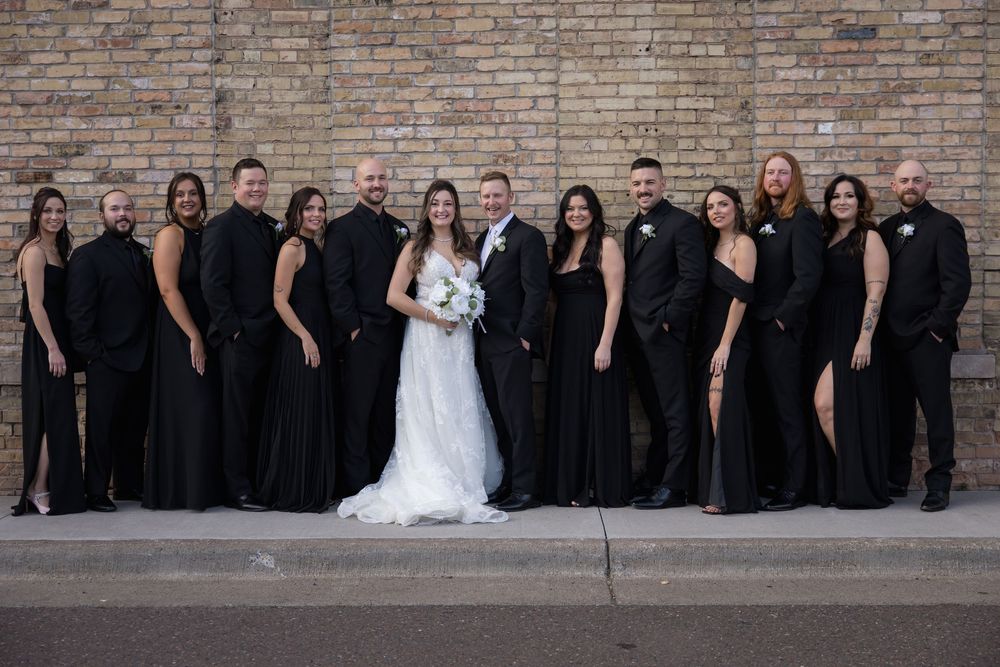 Wedding party in black attire poses with bride in white dress against brick wall.