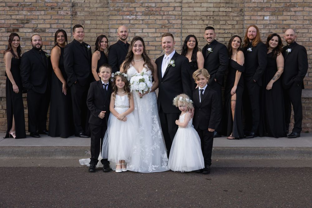 Wedding party in black attire with white gowns stands together for formal portrait against brick wall backdrop.