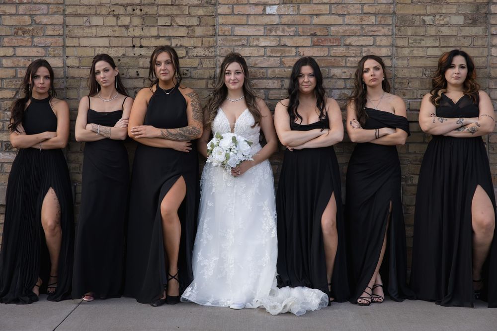 Bridal party with bridesmaids in elegant black dresses poses with bride in white gown against brick backdrop.