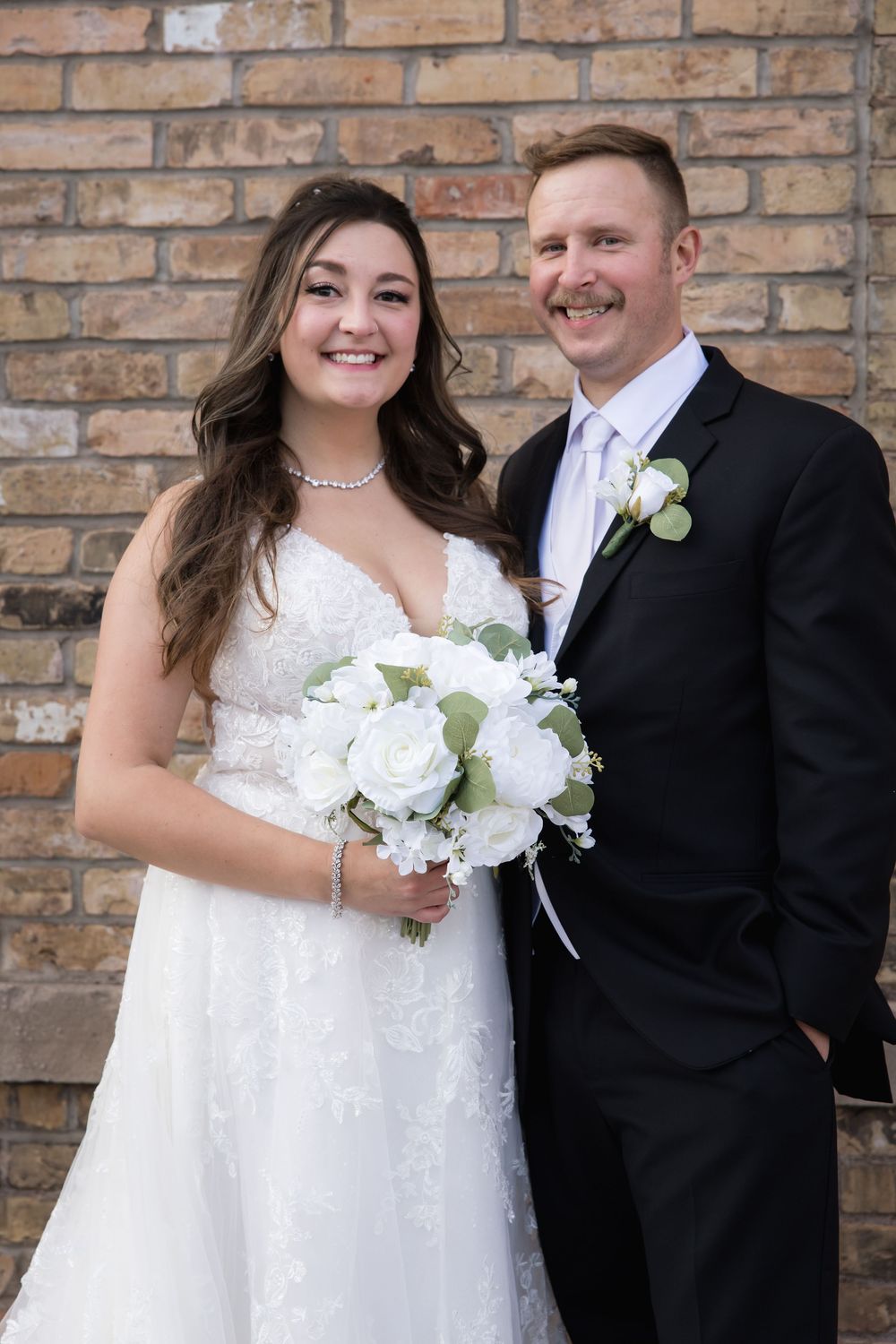 A wedding couple poses against a brick wall backdrop, with the bride holding a white rose bouquet.