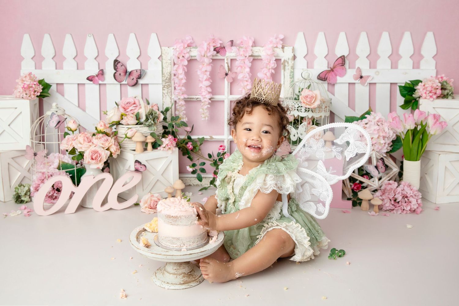 First birthday celebration setup with pink decorations, white picket fence, and cake smash scene.