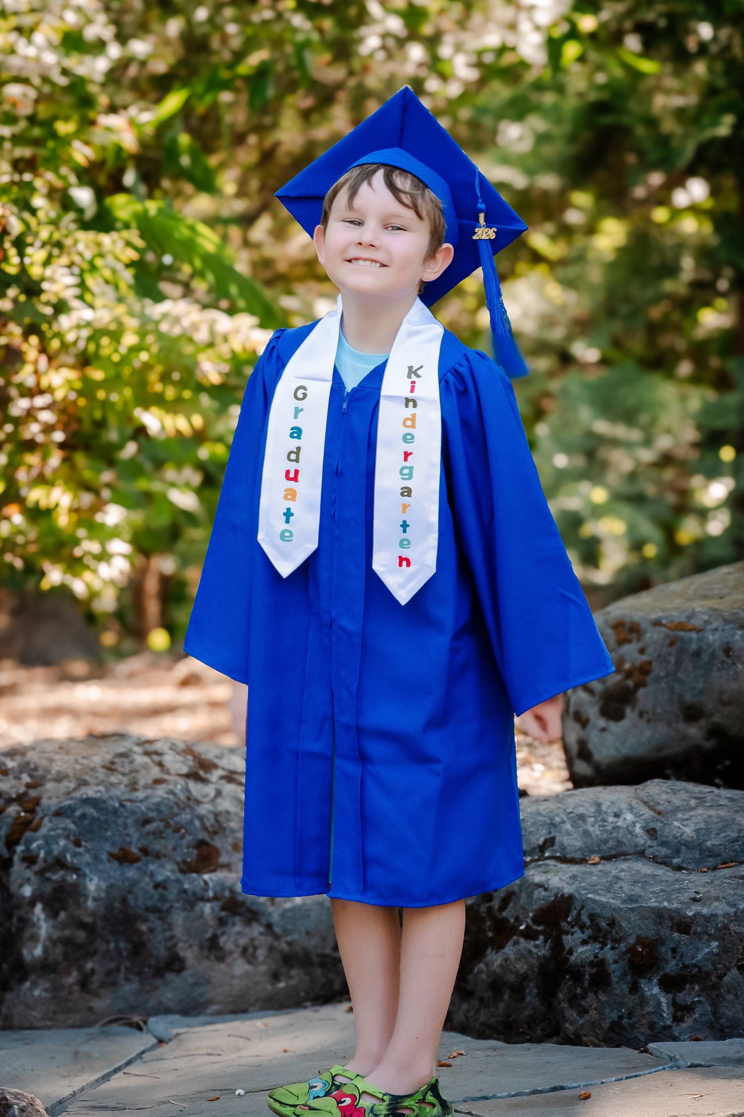 Smiling child in blue kindergarten graduation cap and gown with graduate sash, standing outdoors on stone steps.