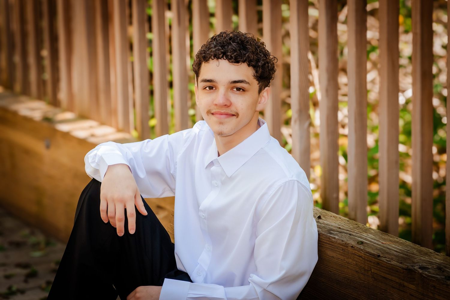 Young man in white dress shirt sitting outdoors by wooden fence, smiling for senior portrait photo.