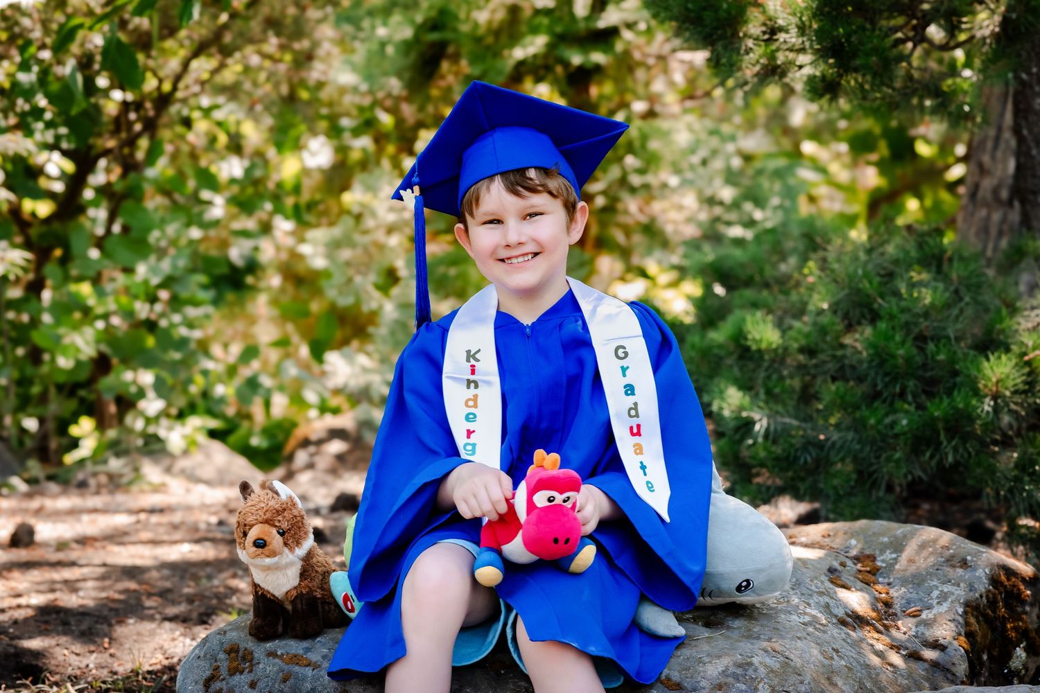 Young child in blue graduation cap and gown holding a red plush toy, sitting on rocks outdoors.