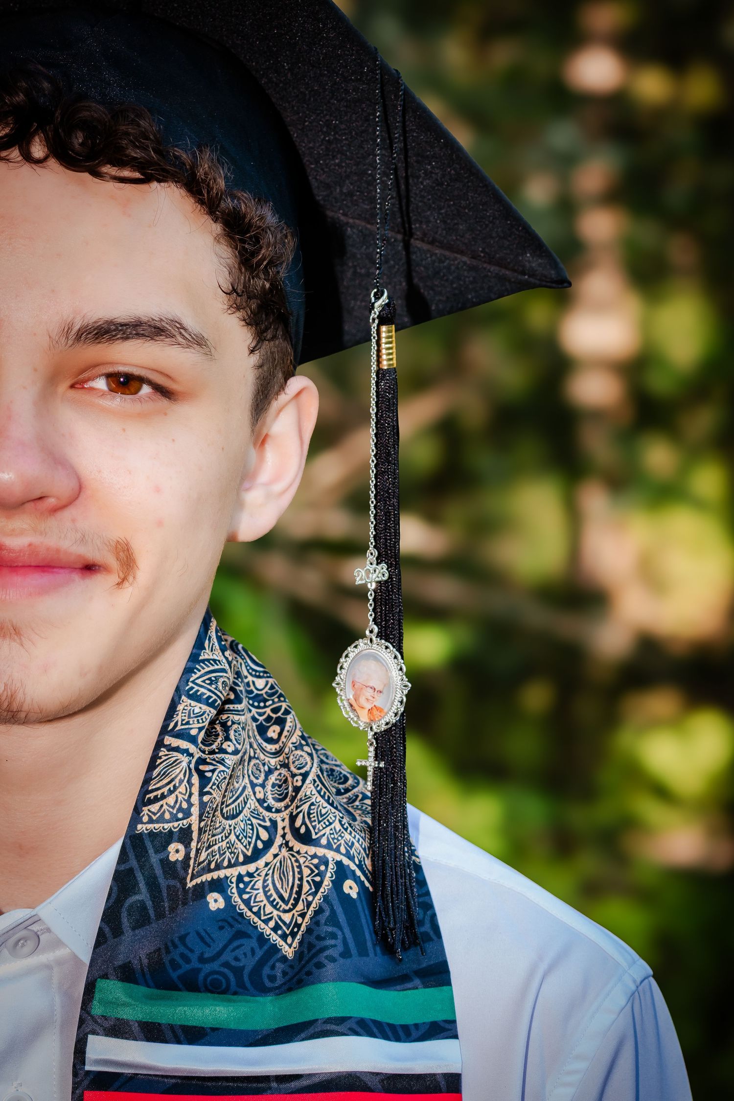 Young man in graduation cap and decorative stole with memorial tassel charm, smiling outdoors.