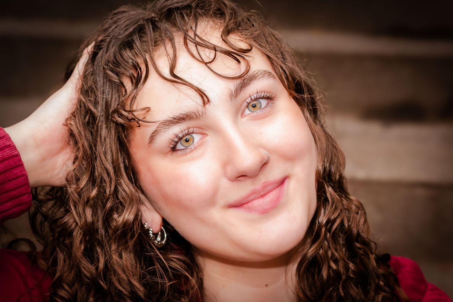 Young woman with curly brown hair smiling, wearing a red top, posing with hand in hair.