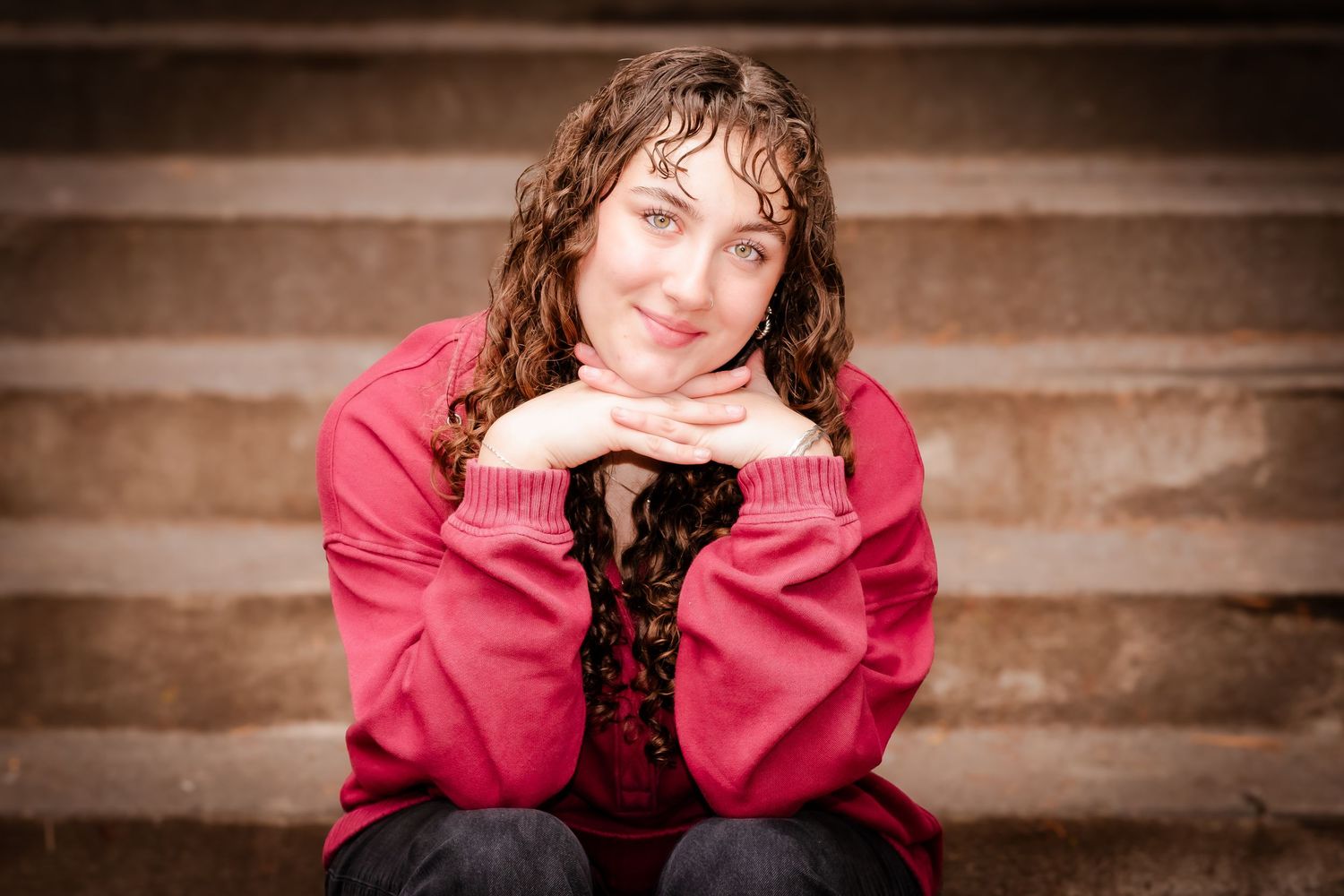 Young woman with curly hair in a red sweater sits on stone steps, smiling with hands clasped under chin.