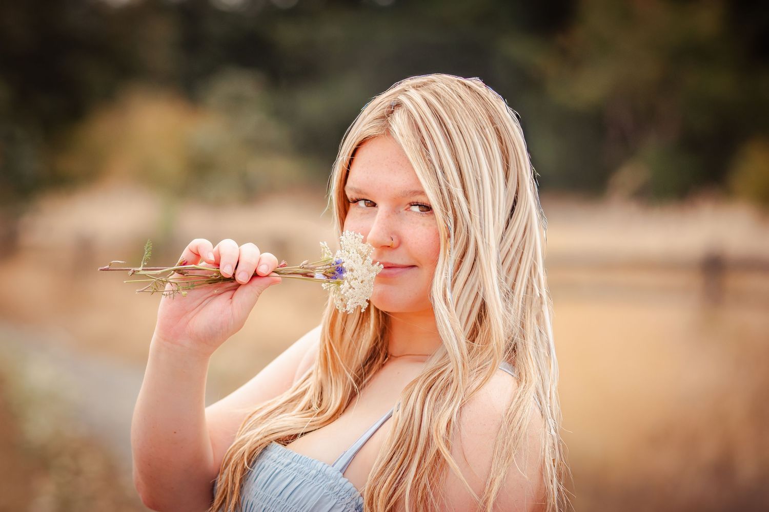 A person with long blonde hair poses against a natural background with golden sunlit tones and dreamy bokeh effect.