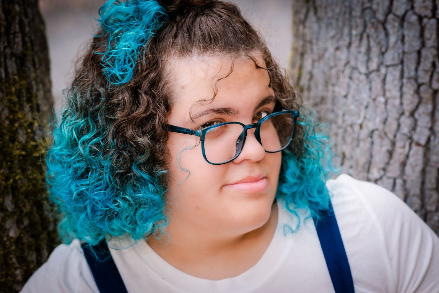 Young woman with curly teal-dyed hair and black glasses posing outdoors near a tree trunk.