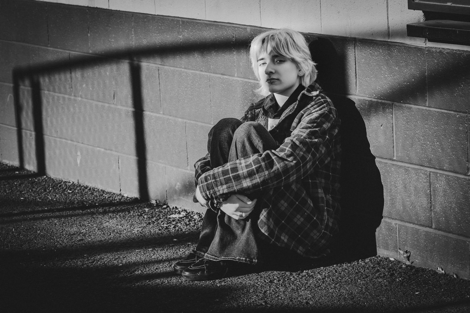 Black and white photo of a young person sitting alone against a concrete wall, hugging their knees in a somber pose.