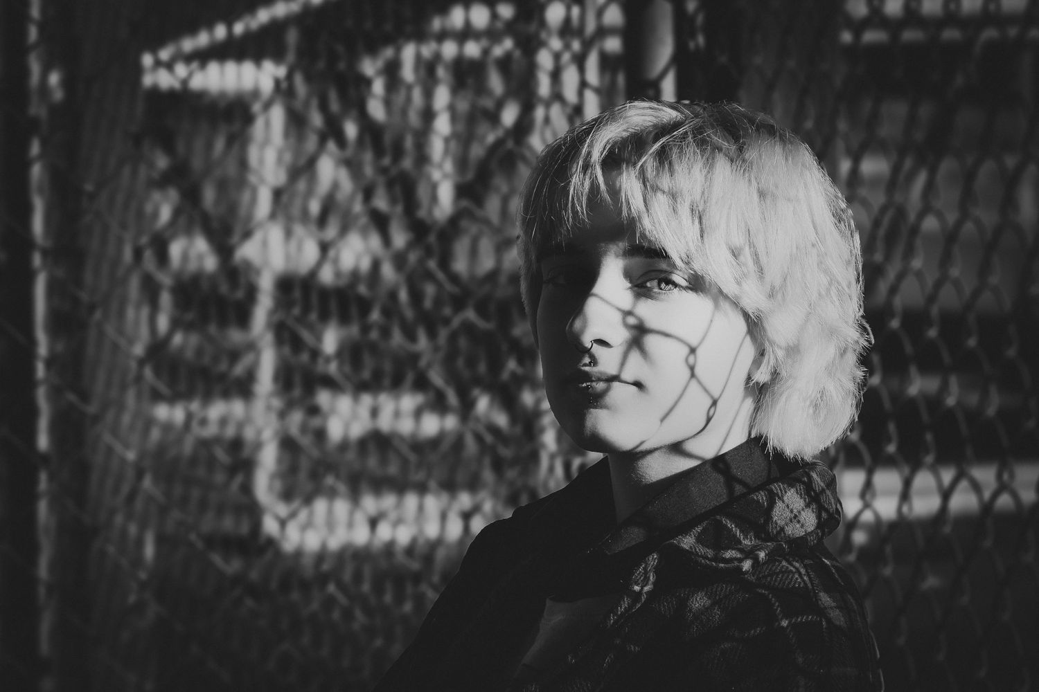 Black and white portrait of a young blonde woman standing in front of a chain-link fence.