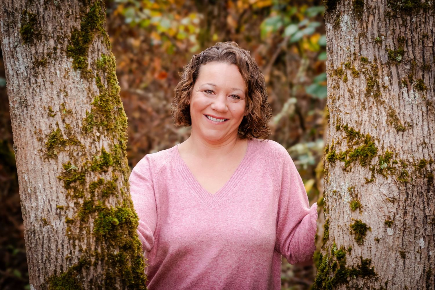 Woman in pink sweater smiling between moss-covered trees in an autumn forest setting.