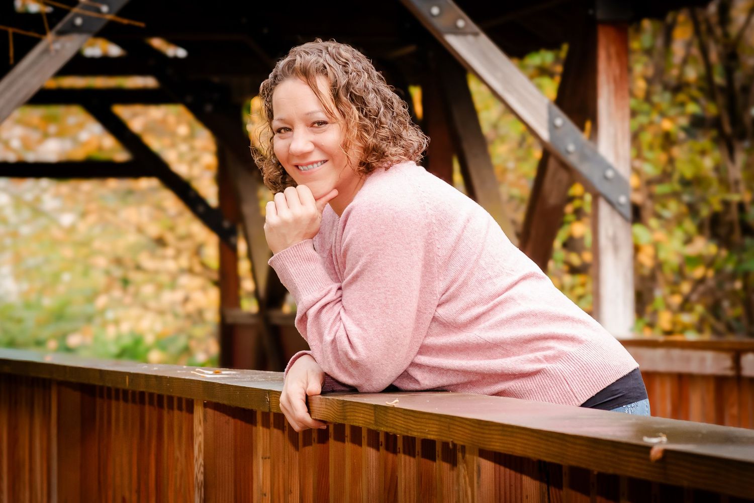 A smiling woman in a pink sweater leans on a wooden railing surrounded by autumn foliage.