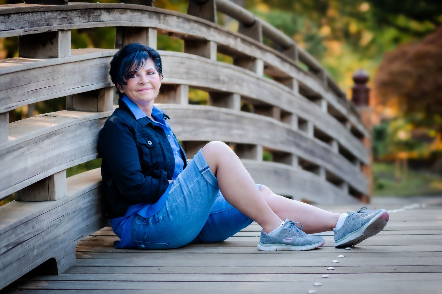 Woman with short dark hair sitting on a wooden bridge, wearing a blue denim jacket and skirt with gray sneakers.