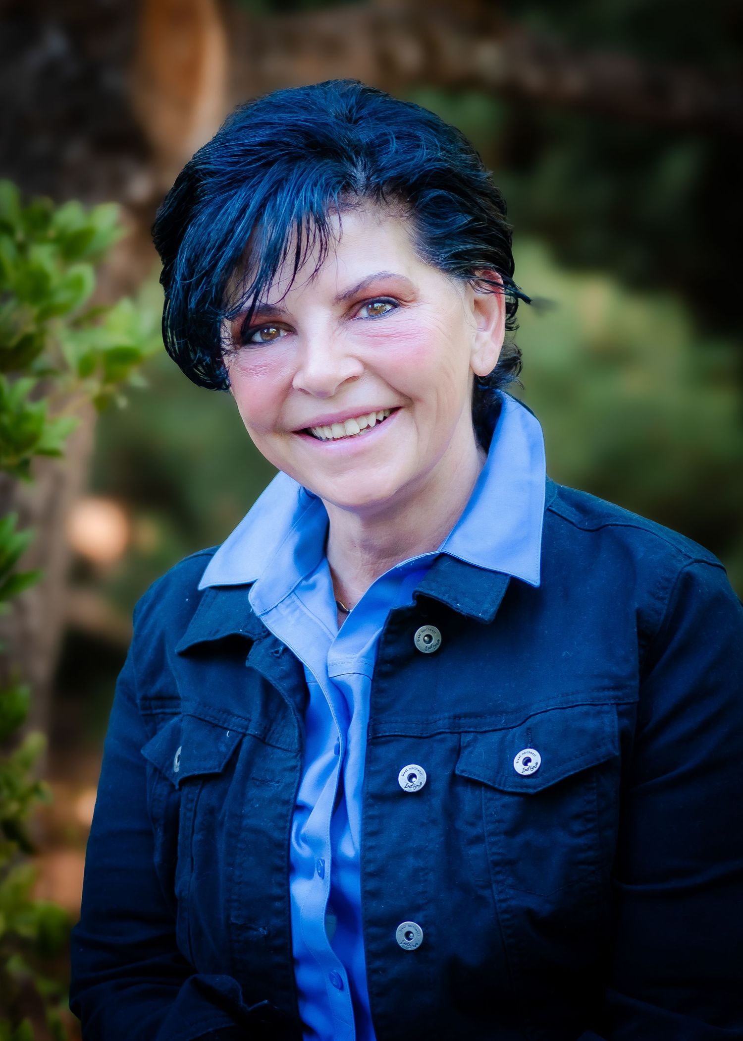 Smiling woman with dark hair wearing a blue denim jacket outdoors in a natural setting.