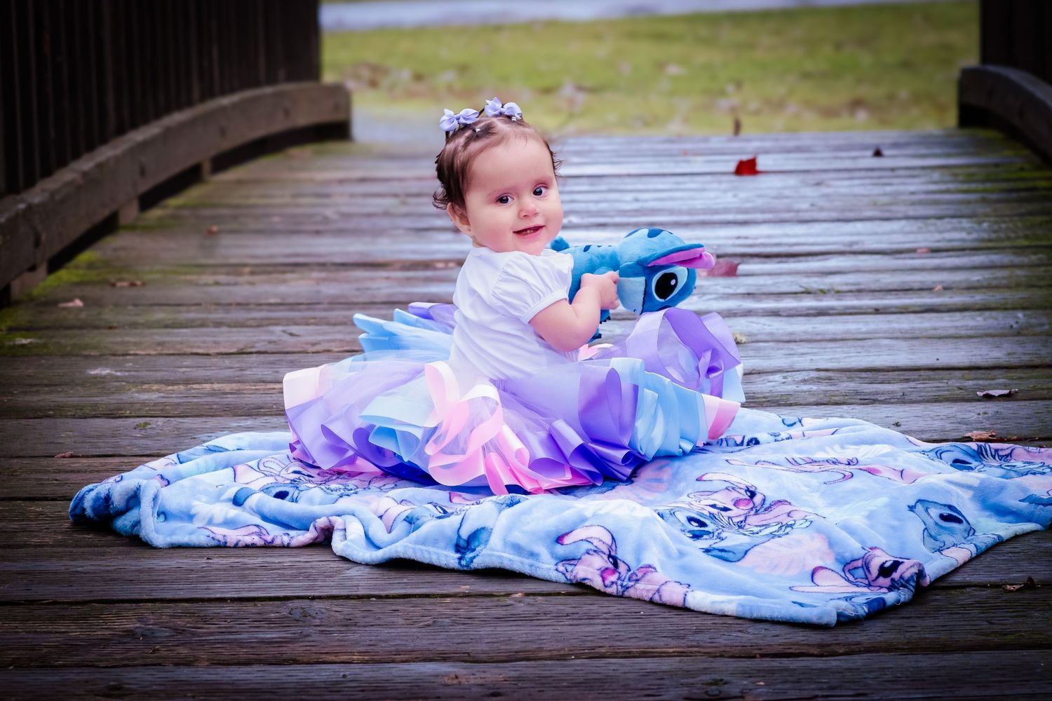 Baby girl in white tutu dress sits on blue blanket on wooden deck holding colorful toy unicorn.