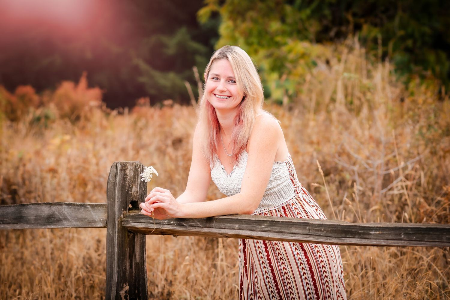 Smiling blonde woman in floral dress leaning on rustic wooden fence in golden field during sunset.