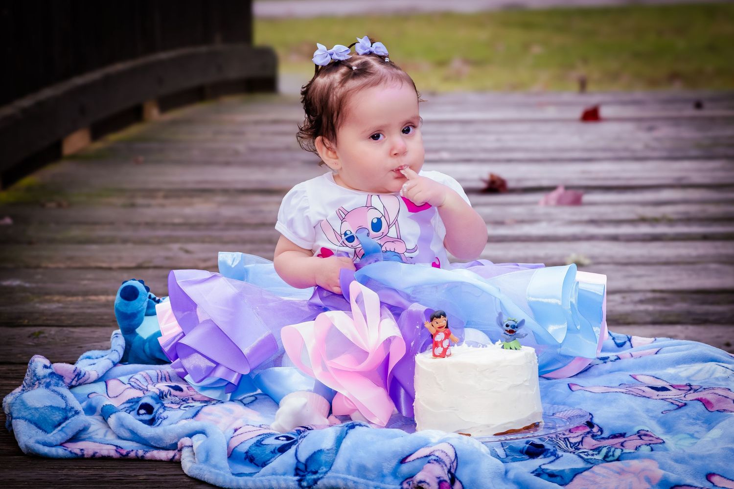 Baby girl in blue tutu sits on wooden deck beside small white cake with Lilo and Stitch themed decorations.