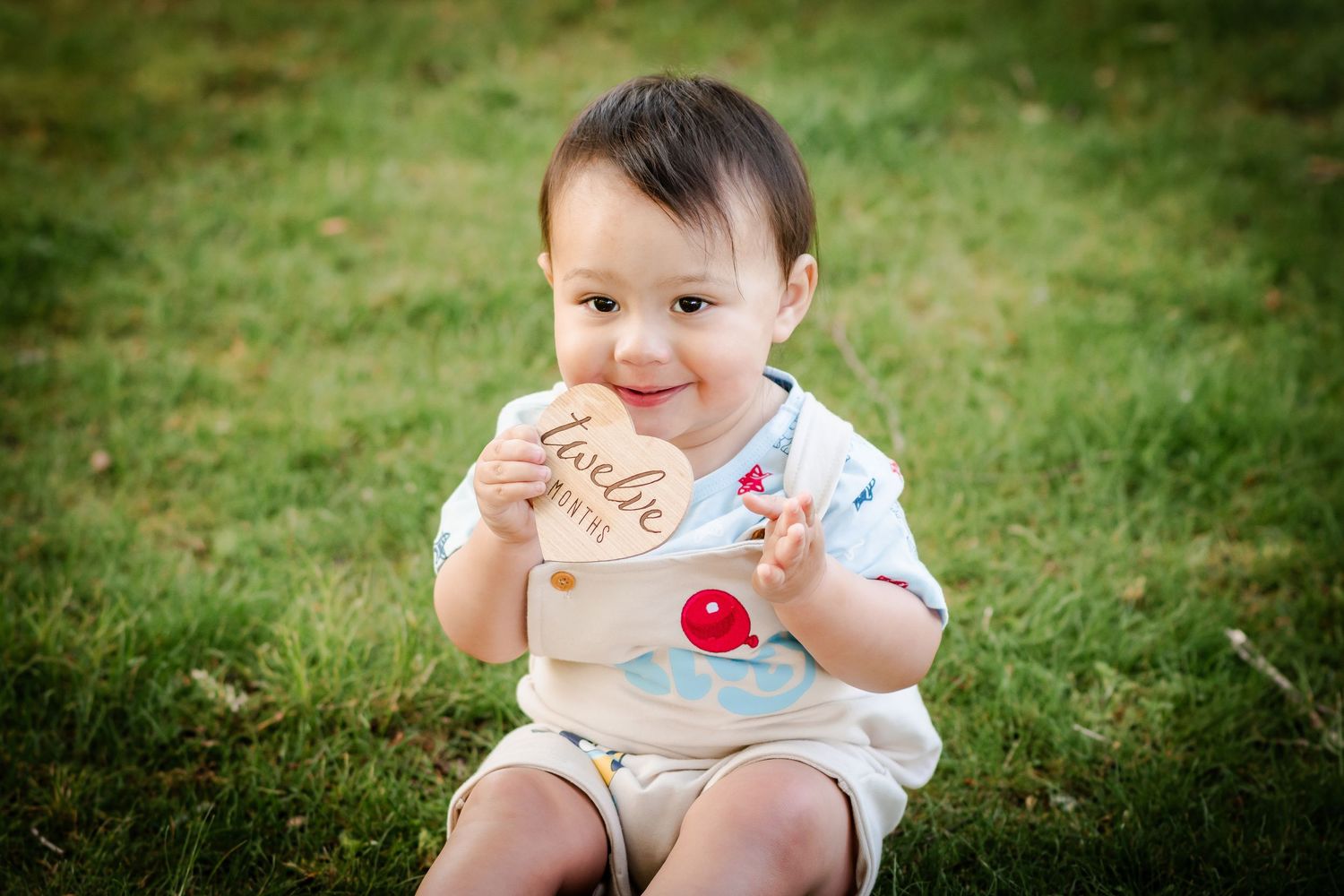 Toddler in white outfit smiles while holding treats on grassy lawn.